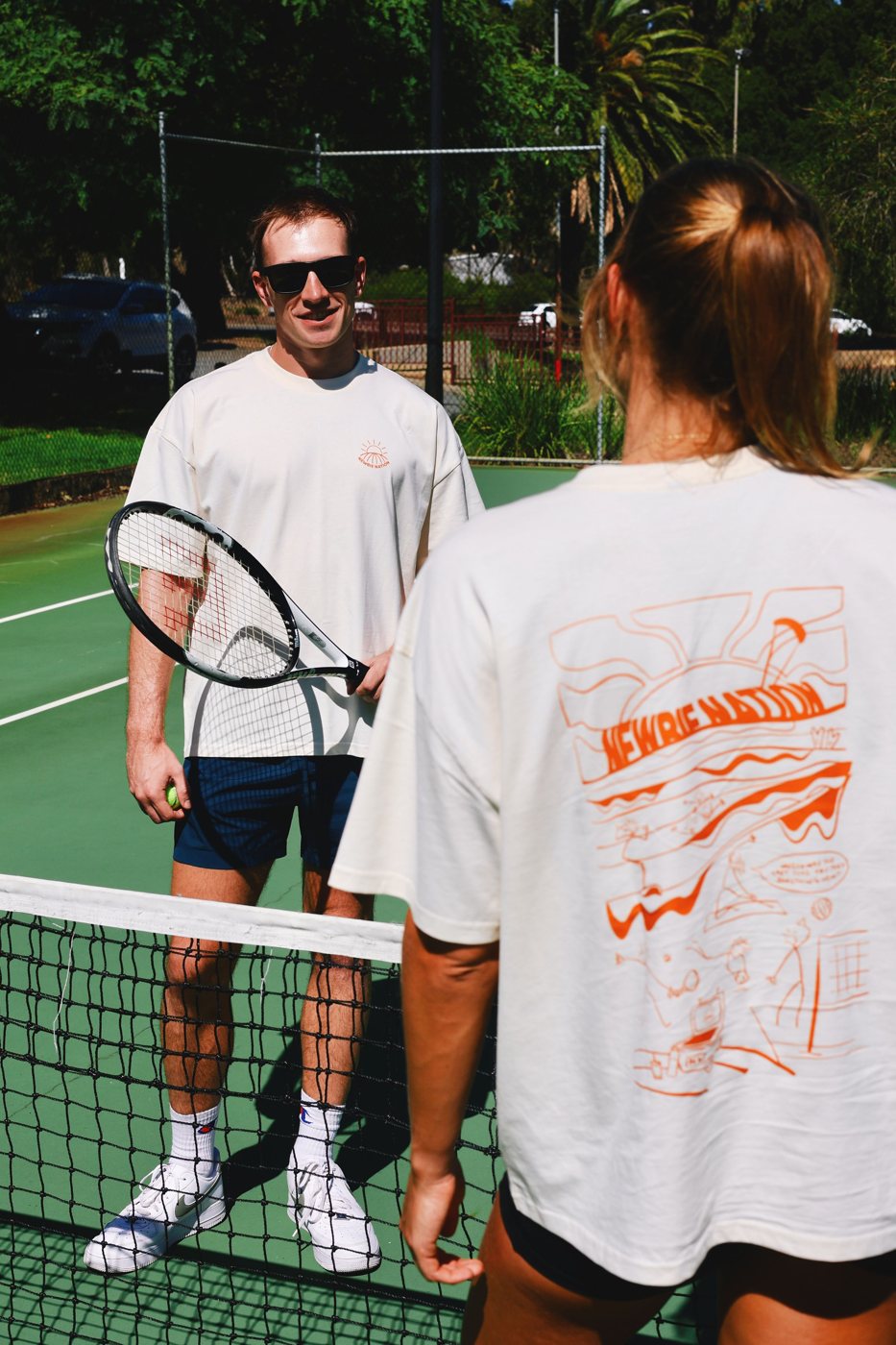 Two newbies on a tennis court wearing the Try Something New Tee, front and back