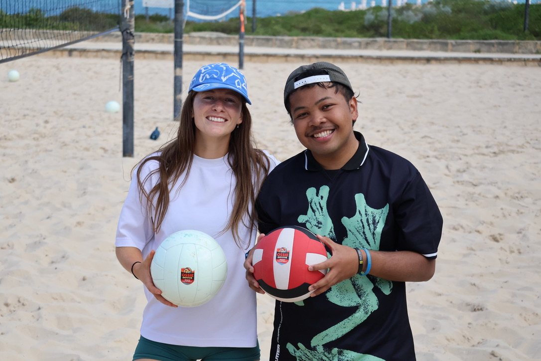 Two newbies smiling with volleyballs on the sand at a beach volleyball social